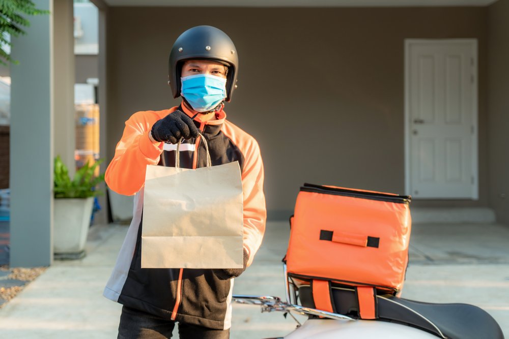 Um motoboy usando um capacete preto, máscara azul, luvas pretas e uma blusa laranja segura para a câmera uma sacola de papel. Ao fundo está sua moto, com uma mochila de entregas laranja, em frente a uma casa de parede marrom e uma porta branca.
