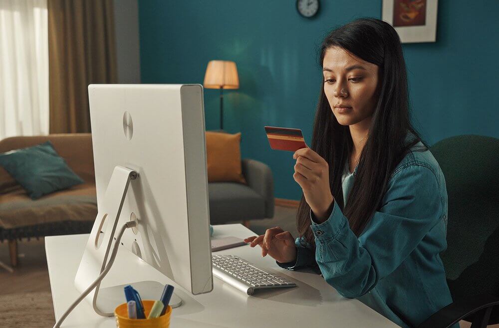 Fundo de uma sala com parede azul. Uma mulher de cabelos longos e escuros sentada em frente a um computador segurando um cartão de crédito em uma das mãos.