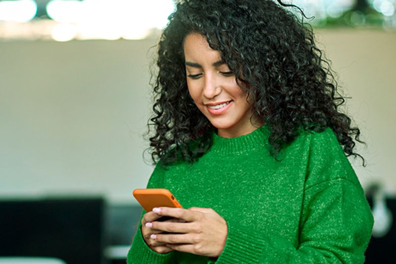 Mulher de cabelos cacheados e blusa verde olha para um celular com uma capa laranja enquanto o segura com as duas mãos
