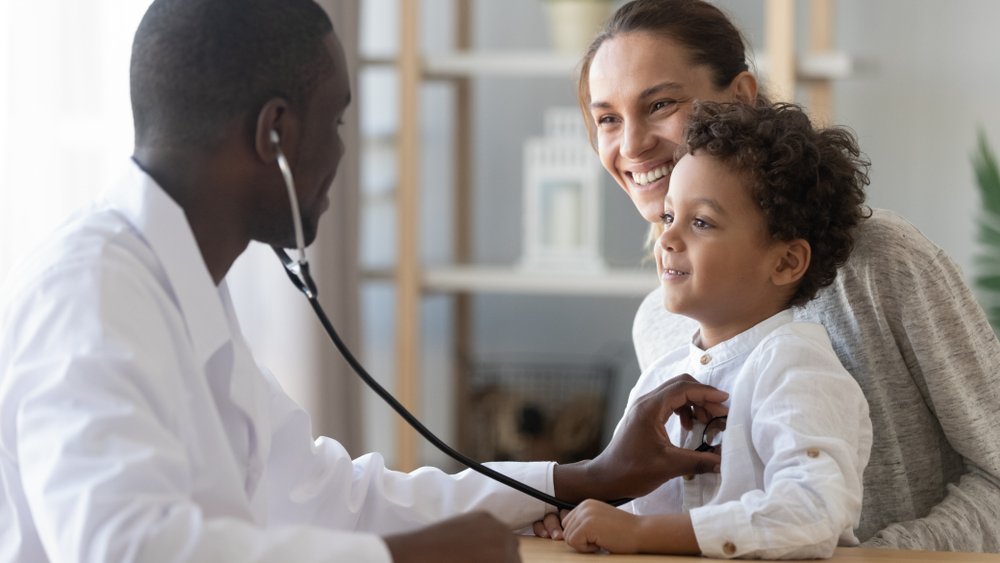 Mãe com uma criança de blusa branca no colo, enquanto um médico negro escuta o coração da criança com um estetoscópio