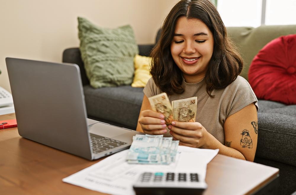 Fundo de uma sala. Mulher sentada na frente de um computador, contando cédulas de dinheiro.